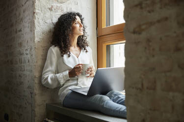 Thoughtful female professional with coffee cup and laptop sitting on window sill - RBF08250