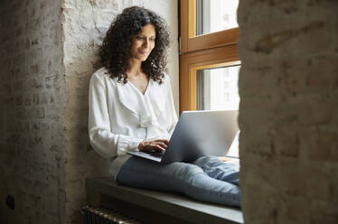 Smiling businesswoman using laptop while sitting on window sill at office - RBF08251