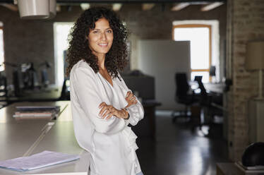 Smiling businesswoman with arms crossed leaning on desk in office - RBF08259