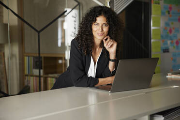 Smiling businesswoman with hand on chin leaning by laptop at workplace - RBF08260