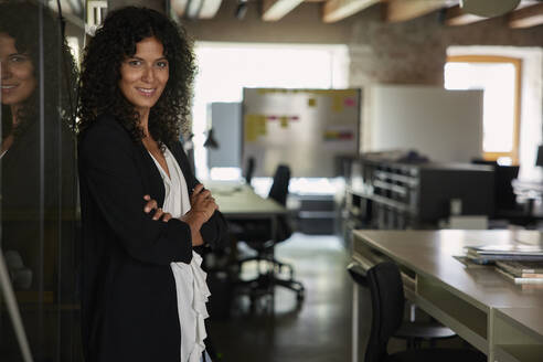 Smiling businesswoman with arms crossed leaning on glass at workplace - RBF08265