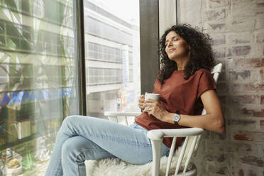 Entspannte Geschäftsfrau, die eine Kaffeetasse hält und mit geschlossenen Augen auf einem Stuhl am Fenster sitzt - RBF08266