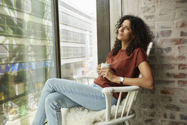 Nachdenkliche Frau mit Kaffeetasse auf einem Stuhl am Fenster sitzend - RBF08267