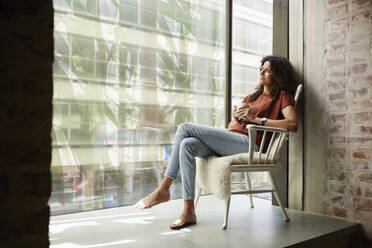 Weibliche Fachkraft hält Kaffeetasse, während sie mit gekreuzten Beinen auf einem Stuhl sitzt - RBF08269