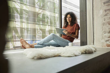 Lächelnde Frau, die ein digitales Tablet benutzt, während sie am Fenster sitzt - RBF08272