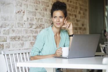 Nachdenkliche Frau sitzt mit Kaffeetasse und Laptop am Schreibtisch am Arbeitsplatz - RBF08294