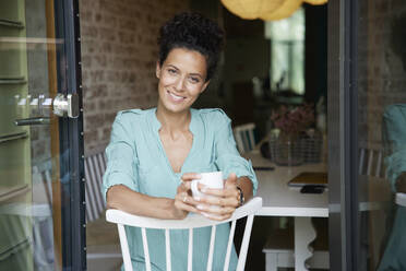 Lächelnde Geschäftsfrau, die eine Kaffeetasse hält und auf einem Stuhl sitzt - RBF08298