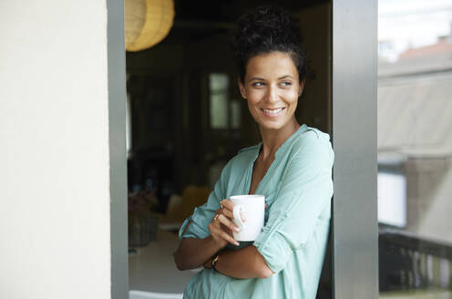 Lächelnde weibliche Fachkraft, die eine Kaffeetasse hält und sich an die Tür lehnt - RBF08301