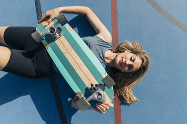 Blond woman with skateboard lying at basketball court during sunny day - JRVF01356
