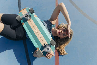 Young woman shielding eyes while lying with skateboard at basketball court - JRVF01357