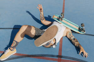 Gefallener junger Mann mit Skateboard im Gesicht auf einem Basketballplatz an einem sonnigen Tag - JRVF01358