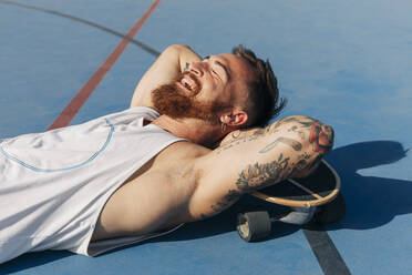Glücklicher junger Mann mit Händen hinter dem Kopf, der sich mit einem Skateboard auf einem Basketballplatz an einem sonnigen Tag ausruht - JRVF01360