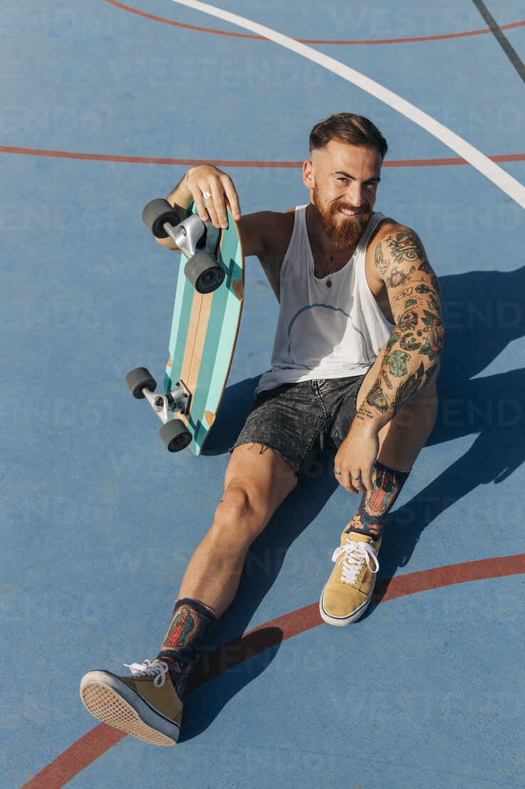 Smiling young man sitting with skateboard at basketball court on