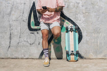 Mann, der auf dem Skateboard eine SMS über sein Smartphone verschickt, während er sich an die Wand lehnt, an einem sonnigen Tag - JRVF01386