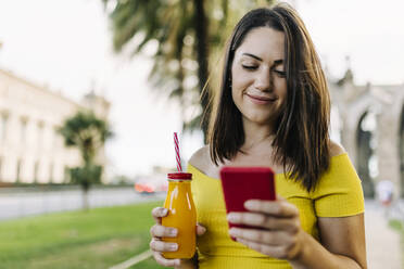 Lächelnde Frau hält eine Saftflasche und benutzt ein Smartphone - XLGF02143