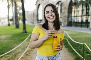 Smiling young woman holding orange juice bottle while standing in park - XLGF02144