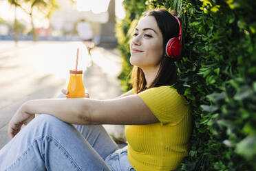 Lächelnde Frau mit Saftflasche hört Musik über Kopfhörer und lehnt sich an eine Hecke - XLGF02160
