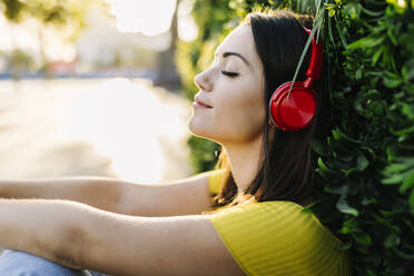 Junge Frau hört Musik über rote Kopfhörer und lehnt sich an eine Hecke - XLGF02162