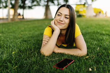 Junge Frau mit Smartphone, die auf dem Rasen liegend nachdenkt - XLGF02176