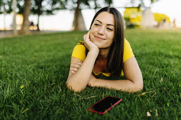 Young woman with smart phone contemplating while lying on lawn - XLGF02176