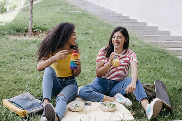 Junge Frau sieht ihre Freundin lachend an, während sie im Park etwas trinkt - TCEF02054
