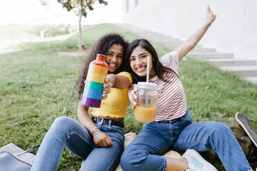 Female friends showing mason jar and bottle while sitting in park - TCEF02058