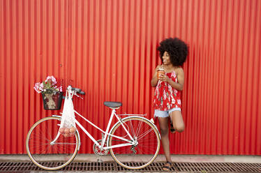 Lächelnde Frau hält einen Smoothie in der Hand, während sie mit dem Fahrrad vor einer roten Wand steht - VEGF04816