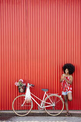 Young woman standing by bicycle in front of red wall - VEGF04817