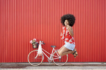Glückliche Afro-Frau, die mit dem Fahrrad vor einer roten Wand springt - VEGF04820