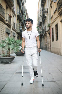 Young man walking in alley with help of crutches amidst buildings - AGOF00191