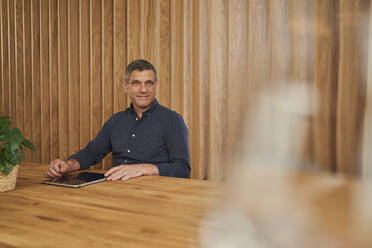 Lächelnde männliche Fachkraft sitzt mit digitalem Tablet am Konferenztisch im Sitzungssaal - AKLF00366