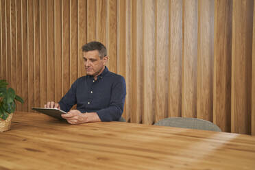 Reifer männlicher Berufstätiger, der ein digitales Tablet benutzt, während er am Konferenztisch sitzt - AKLF00369