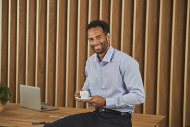 Smiling businessman with coffee cup sitting on conference table - AKLF00377