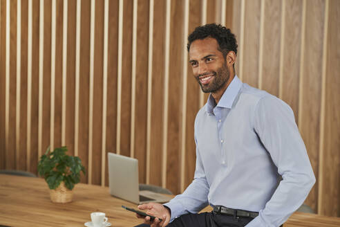 Lächelnder Geschäftsmann, der ein Mobiltelefon hält, während er im Sitzungssaal sitzt - AKLF00379