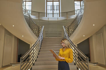 Blonde Frau benutzt ein digitales Tablet, während sie vor einer Treppe steht - AKLF00392