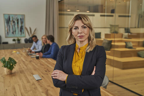 Confident blond businesswoman standing with arms crossed in board room - AKLF00439
