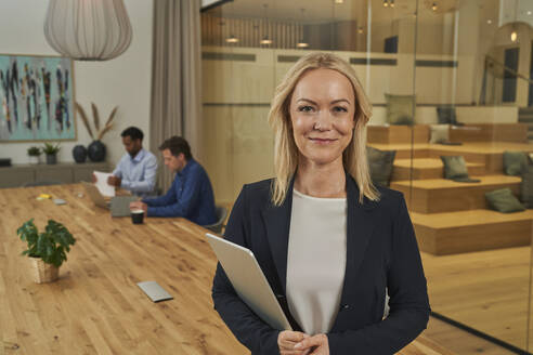 Smiling blond businesswoman holding digital tablet while standing in board room - AKLF00445