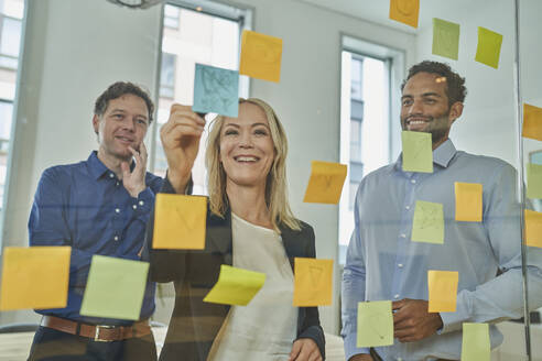 Smiling businesswoman writing on adhesive note while discussing with male colleagues in board room - AKLF00451