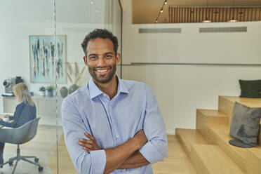 Smiling male professional with arms crossed leaning on glass wall in office - AKLF00462