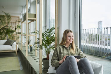 Glückliche Praktikantin mit digitalem Tablet, die durch das Fenster schaut, während sie im Büro sitzt - AKLF00478