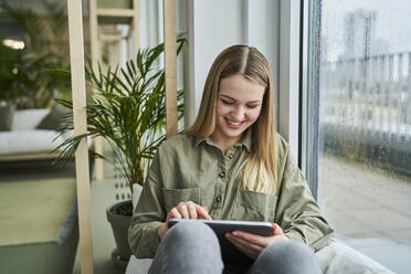 Lächelnde Praktikantin, die ein digitales Tablet benutzt, während sie an einem Glasfenster im Büro sitzt - AKLF00479