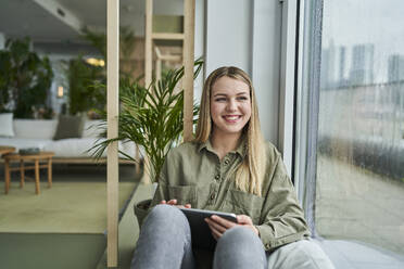 Lächelnde Praktikantin im Teenageralter mit digitalem Tablet, die durch das Fenster schaut, während sie im Büro sitzt - AKLF00481