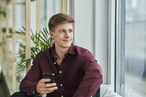 Lächelnder junger Geschäftsmann schaut weg, während er sein Smartphone im Büro hält - AKLF00486