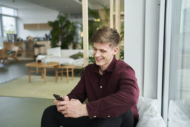 Lächelnder männlicher Berufstätiger, der über sein Smartphone im Büro Textnachrichten verschickt - AKLF00487