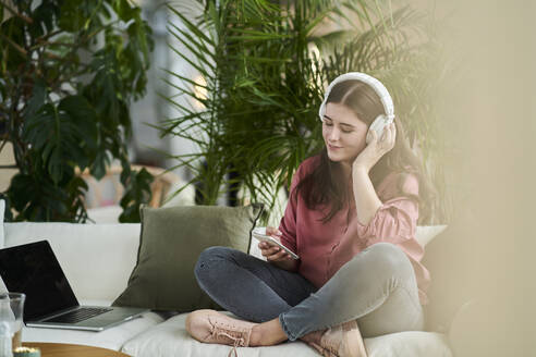 Junge Frau hört Musik über Kopfhörer, während sie ihr Smartphone in der Pause im Büro benutzt - AKLF00492