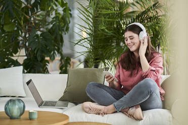 Lächelnde junge Berufstätige, die ein Mobiltelefon benutzen und über Kopfhörer Musik hören, während sie auf dem Sofa im Büro sitzen - AKLF00493