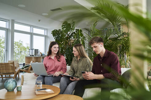 Male professional using smart phone while sitting by female colleagues discussing in office - AKLF00496