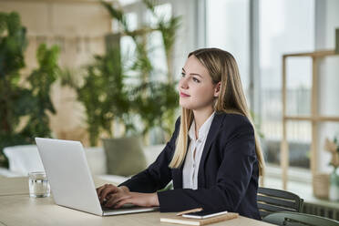 Thoughtful teenage intern looking away while sitting with laptop at desk in office - AKLF00507