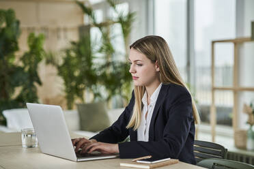 Blond female trainee typing on laptop while sitting at desk in office - AKLF00508