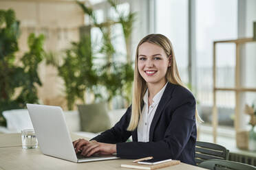 Lächelnde blonde weibliche Auszubildende sitzt mit Laptop am Schreibtisch im Büro - AKLF00510